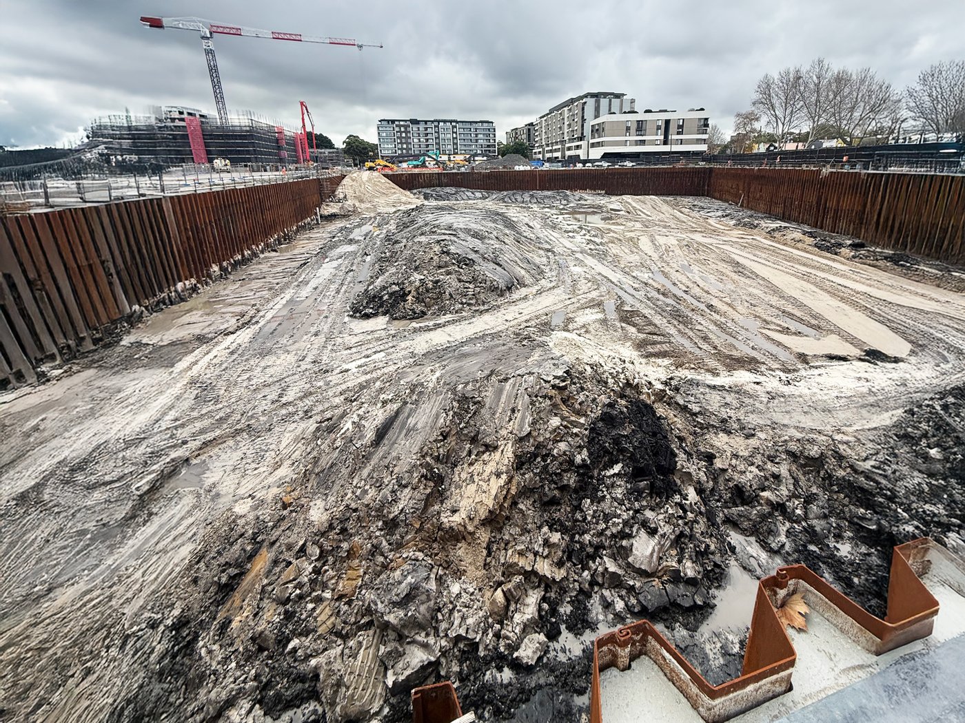 A deep shored excavation on an urban renewal site, cranes and buildings behind
